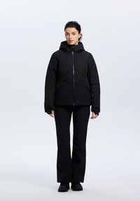 Chaqueta negra aislante con capucha, que presenta un patrón acolchado en la parte superior y una sección inferior suave, combinada con pantalones negros ajustados.
