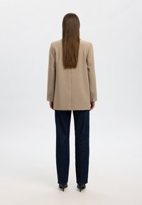Beige blazer with long sleeves and button accents, paired with dark blue straight-leg jeans and black pointed heels, viewed from the back.