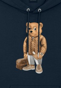 Sudadera de color navy con un gráfico de un oso pardo vestido con un suéter de punto trenzado, sosteniendo una bolsa marrón y una cartera, con un reloj y zapatos de color claro.