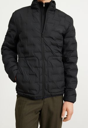 Mann trägt eine schwarze, gesteppten Reißverschlussjacke mit Taschen und Barbour International-Logo auf der Brust, kombiniert mit olivgrünen Hosen.