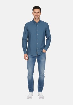 Hombre sonriente de pie, con una camisa azul de botones, jeans azul claro y zapatillas blancas, con una mano en el bolsillo, sobre un fondo liso.