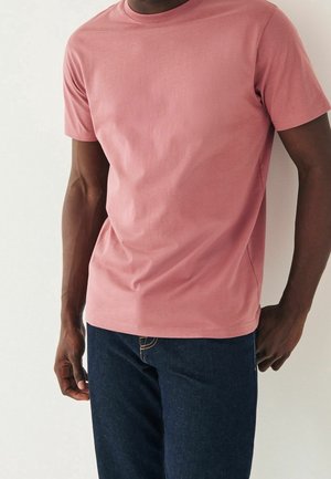 Mann trägt ein kurzärmeliges T-Shirt in staubigem Roseton mit Rundhalsausschnitt und dunkelblaue Jeans, steht vor hellem Hintergrund.