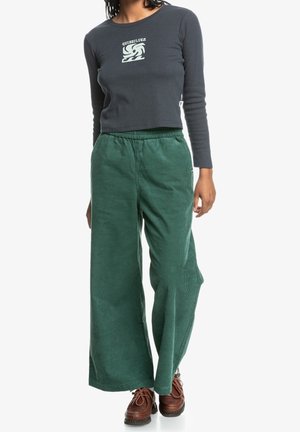 Personne portant un crop top gris foncé à manches longues avec un logo et un pantalon large en velours côtelé vert, associé à des chaussures lacées marron.