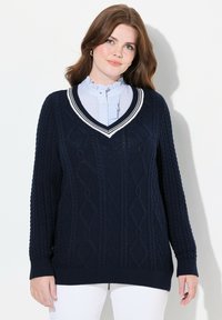 Marinblauer Kabelstrickpullover mit V-Ausschnitt und weißen Streifen. Verfügt über strukturierte Rauten-Muster sowie gerippte Bündchen und Saum. Wird über einem kragenlosen Hemd getragen.