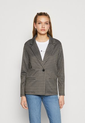Kostkovaný blazer v černé a béžové barvě, s jedním knoflíkem, se zářezovými klopami, dvěma předními kapsami, nošený přes bílé tričko a modré džíny.