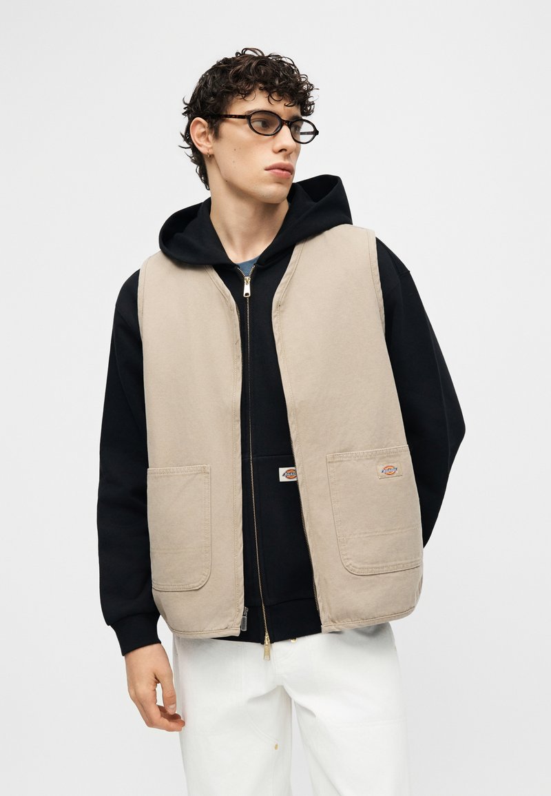 Joven con cabello rizado que lleva gafas con montura carey, un chaleco beige sobre una sudadera negra y pantalones blancos, mirando hacia un lado.