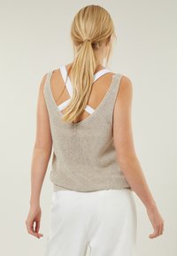 Débardeur beige tricoté avec un dos en V, équipé de larges bretelles et d'une coupe ample. Porté sur un pantalon blanc, mettant en valeur un motif texturé.