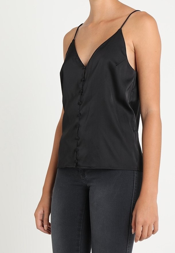 Top sin mangas de satén negro con tirantes finos y botones cubiertos de tela en el frente, combinado con jeans gris oscuro, en una persona de pie.