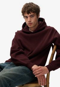 Sudadera burdeos con una Capucha grande y relajada, textura suave y puños de punto, combinada con unos jeans de corte holgado en azul claro. Sentado en una silla de madera.