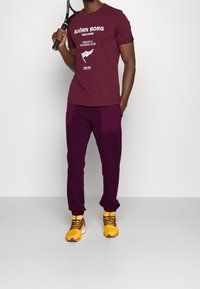 Bordeauxrotes T-Shirt mit weißem Druck und lila Trainingsthose. Gelbe Sneakers mit schwarzen und roten Schnürsenkeln. Hält einen Tennisschläger.