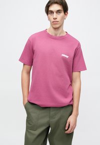 Růžové bavlněné tričko s kulatým výstřihem a krátkými rukávy, na hrudi se bílým logem "obey". Spárované s olivově zelenými kalhotami.