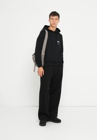 Sweat-shirt à capuche noir avec poche frontale, associé à un pantalon noir ample et des baskets noires. Sac à épaule gris en bandoulière.
