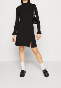 Musta, ribsikuvioinen, pitkähihaisen mekko, jossa on korkea kaulus ja etureikä. Yhdistetty mustien loafereiden ja valkoisten nilkkasukien kanssa sekä mustan laukun kanssa.