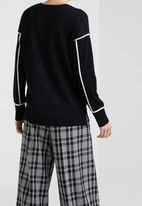 Schwarzer Pullover mit weißen Akzenten entlang der Ärmel, gerippter Saum und Bündchen, kombiniert mit weit geschnittenen schwarz-weißen Karohosen.