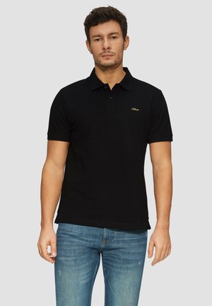 Schwarzes Polohemd aus Baumwolle mit klassischem Kragen, zwei Knöpfen und einem kleinen goldenen Logo auf der linken Brust. Getragen mit blauen Jeans.