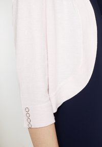 Ljusrosa kofta med mjuk stickad textur, med en rundad fåll och tre metallknappar på ärmslutet. Bärs över ett mörkt plagg.