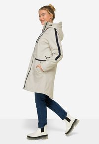 Parka impermeabile beige chiaro con cappuccio, strisce nere lungo le maniche, tasche con zip e vestibilità rilassata, indossata sopra jeans blu e stivali bianchi.