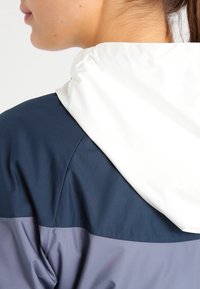 Färgblockerad jacka med en vit huva, mörkblå överdel och grå underdel, tillverkad av ett mjukt och lätt material.