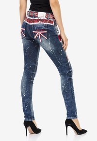 Schmale Jeans in Blau mit weißem Farbspritzer, akzentuierte Taschen mit Union Jack Design, verzierten Bund und High Heels.