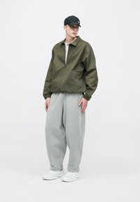 Jopič z zeleno olivno zadrgo, sive široke trenirke, bele superge in črna kapa. Jopič iz gladkega blaga z elastičnimi manšetami.