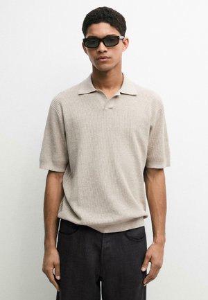 Uomo che indossa occhiali da sole scuri, una polo beige a maniche corte in maglia e pantaloni scuri, in piedi davanti a uno sfondo bianco semplice.