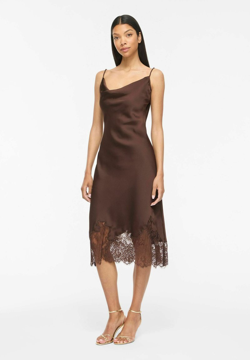 Femme aux longs cheveux noirs porte une robe en satin marron sans manches avec un ourlet en dentelle et des talons dorés à brides, debout devant un fond blanc uni.