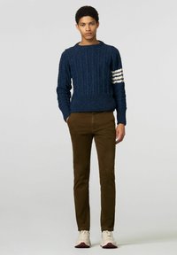 Marineblauwe gebreide trui met ribbelstructuur en witte gestreepte accenten op de linkermouw, in combinatie met bruine slim-fit broek en sneakers.