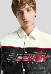 Jeune homme aux cheveux bouclés portant une veste en cuir noir et blanc avec des accents rouges et le logo "Équipe Sportive Redway", fond neutre.