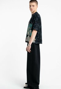 Młody mężczyzna stojący bokiem, ubrany w czarną koszulkę z grafiką przedstawiającą tygrysa, szerokie czarne spodnie, zegarek i czarne buty na białym tle.