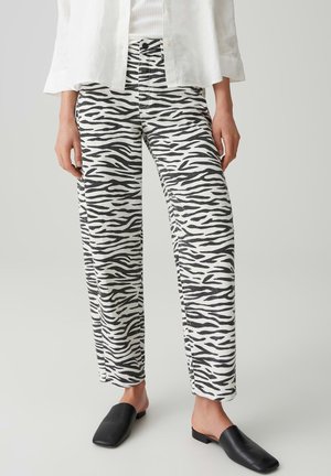 Person trägt schwarz-weiß gestreifte Zebrahosen, weißes Hemd und schwarze Slipper, steht vor einem einfarbigen hellen Hintergrund.