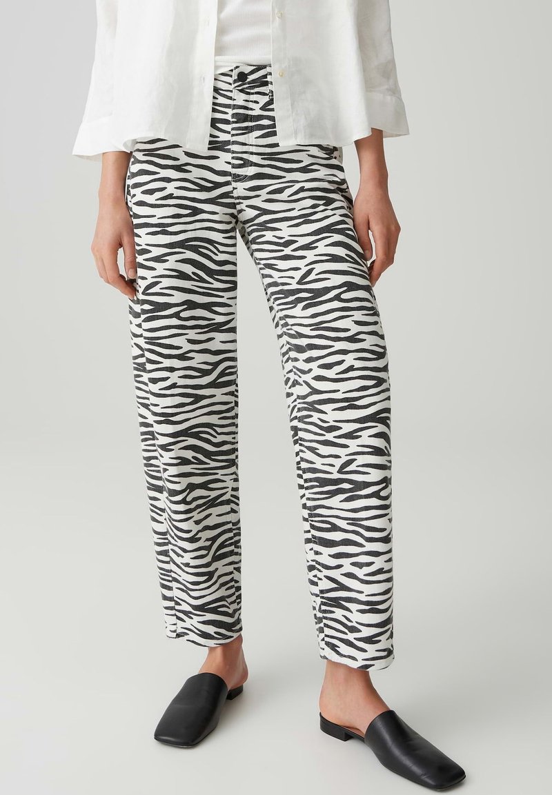 Person trägt schwarz-weiß gestreifte Zebrahosen, weißes Hemd und schwarze Slipper, steht vor einem einfarbigen hellen Hintergrund.