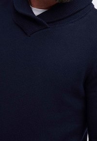 Pull en tricot bleu marine avec un col châle, présentant un tissage texturé. Détails comprenant des poignets et un ourlet côtelés avec une légère brillance.