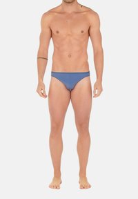 Männlicher Oberkörper und Beine in blauen Badehosen, barfuß stehend vor einem schlichten hellen Hintergrund.