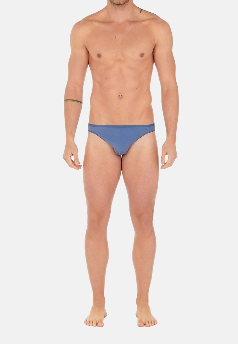 Männlicher Oberkörper und Beine in blauen Badehosen, barfuß stehend vor einem schlichten hellen Hintergrund.