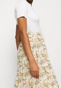 Kukallinen mekko, jossa valkoinen neulottu yläosa ja laskeutuva helma pastellivärisine kukkakuoseineen. Kangas on pehmeä ja siinä on kerroksittainen muotoilu.