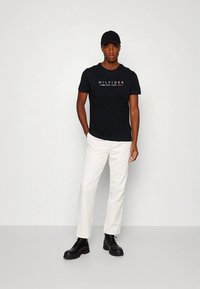 Camiseta negra de algodón con texto blanco que dice "HILFIGER NEW YORK", combinada con pantalones blancos y botas negras con cordones. Modelo de pie contra un fondo liso.
