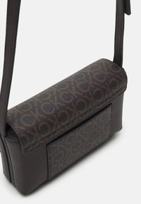 Pieni tummanruskea olkapussukka, jossa on säädettävä hihna, teksturoitu läppä ja tasku, jossa on abstrakti CK-logo-kuvio oranssina.