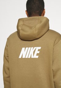 Sudadera con capucha marrón de tela texturizada. Presenta un logo blanco destacado de "NIKE" en la parte trasera, con capucha ajustable con cordón y un ajuste relajado.