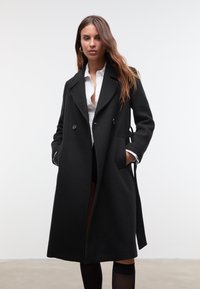 Manteau en laine noir avec un design à double boutonnage, des revers crantés, des poches latérales et une taille à nouer. Présente une texture lisse et une silhouette structurée.
