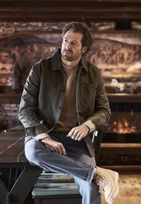 Olivgrüne Jacke über einem hellbraunen Pullover, mit blauen Jeans und hellbraunen Stiefeln. Die Jacke hat eine Brusttasche und Knopfdetails.