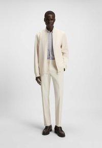 Bomber jacket beige chiaro sopra una camicia a fantasia, abbinato a pantaloni coordinati e scarpe marroni scure. Texture lisce, design minimalista.