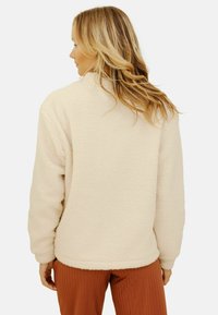 Pull en polaire crème avec manches longues, col rond et finition texturée, associé à un pantalon côtelé couleur rouille. Vue de dos présentée.