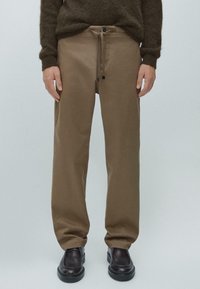 Brązowe bawełniane spodnie o luźnym kroju, z sznurkiem w pasie i zapięciem na guziki, w połączeniu z czarnymi skórzanymi butami. Gładka faktura, minimalistyczny design.