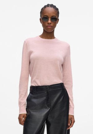 Femme aux cheveux tressés portant des lunettes de soleil rondes noires, un pull rose clair à manches longues et un pantalon en cuir noir taille haute.