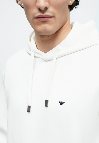 Baltas džemperis su gobtuvu, priekyje kangaroo tipo kišenė, su raišteliais, kurių galiukai yra metaliniai pilkos spalvos, ir nedideliu tamsiai mėlynu logotipu kairėje krūtinės pusėje.