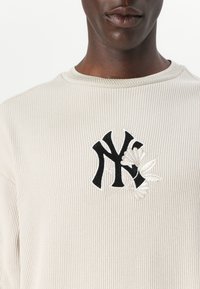 Krem fôret ribbet sweatshirt med en svart NY-logo med blomstermønster. Rund halslinning og teksturert stoff. Nærbilde av overkroppen.