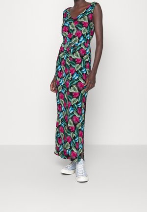 Femme portant une robe sans manches, longue jusqu'aux chevilles, à imprimé tropical avec des feuilles vertes, roses et bleues, associée à des baskets bleu clair.