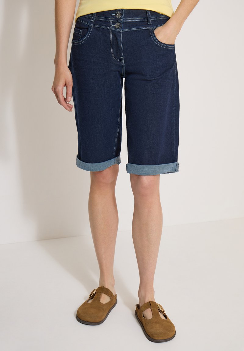 Personne portant un short en denim bleu foncé retroussé, un haut jaune et des chaussures marron à enfiler, debout avec une main dans la poche.