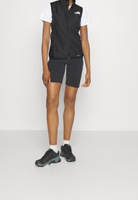 Svart ärmlös väst över en vit t-shirt med kort ärm, parat med svarta shorts och svarta vandringsskor med teal-accenter; vita strumpor.