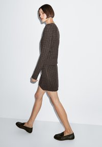 Brauner Strickpullover und passender Rock mit gerippter Struktur, kombiniert mit dunklen Loafers, vor schlichtem weißem Hintergrund präsentiert.